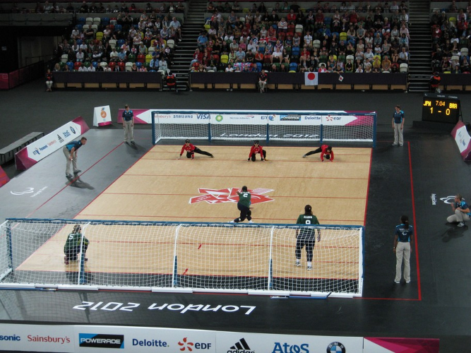 Die Aufnahme zeigt eine Szene aus einem Goalball-Spiel der brasilianischen und der japanischen Frauenteams. Aus einer erhöhten Perspektive hinter dem Tor der Brasilianerinnen ist der Wurf der mittleren der drei Sportlerinnen in grün-schwarzer Kleidung zu sehen. Während die brasilianische  Spielerin gerade den Ball in Richtung japanisches Tor rollt, versuchen die drei Gegnerinnen in rot mit ihrer Körperfläche das eigene Tor zu verteidigen, das sich über die gesamte, neun Meter breite Spielfläche erstreckt. Das Publikum in der voll besetzten Halle ist im Hintergrund zu erkennen.