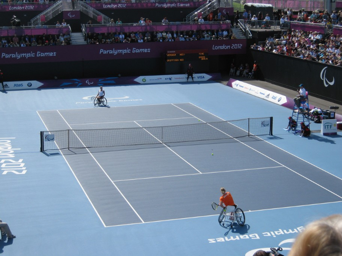 Aus einer erhöhten Position schräg hinter dem Tennisfeld sind zwei Rollstuhltennisspielerinnen beim Einschlagen vor dem Match zu erkennen. Die gut zu erkennende Punkteanzeige verrät, dass es sich um zwei Niederländerinnen handelt. Der Bildvordergrund zeigt eine orange gekleidete Spielerin beim Ausschwung der Schlagbewegung, auf der anderen Seite des Netzes erwartet ihre weiß gekleidete Kontrahentin den Ball. Um das Spielfeld sind die voll besetzten Tribünen und mit Werbung versehenen Banden zu sehen.