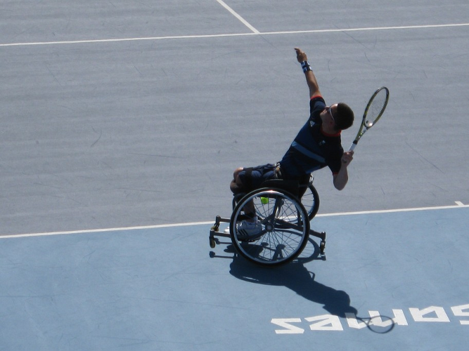 Das Bild zeigt einen Rollstuhltennisspieler in Großaufnahme aus einer erhöhten Perspektive hinter dem Spielfeld. Der Linkshänder holt gerade zum Aufschlag über Kopf aus und stabilisiert die Bewegung aus dem Oberkörper mit dem erhobenen rechten Arm. Durch die starke Sonneneinstrahlung hebt sich der Spieler in dunkler Kleidung als Silhouette vom hellblauen Boden des Tennisfeldes ab. 