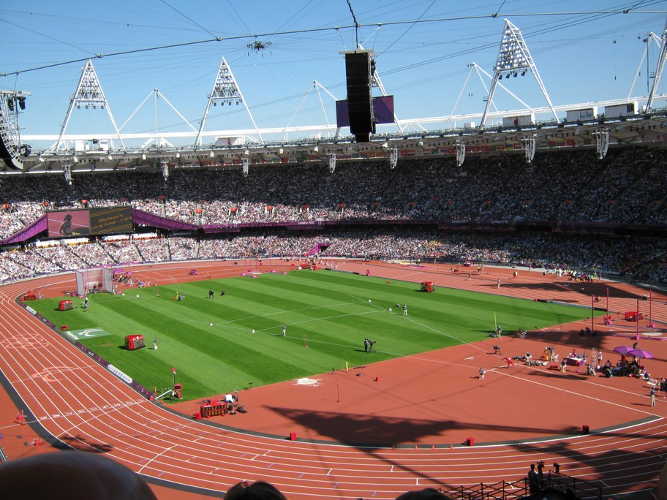 Die Aufnahme zeigt von einem hohen Tribünenplatz aus das weite Rund des Londoner Olympiastadions. In der ausverkauften Arena finden gerade mehrere Leichtathletikwettkämpfe statt, die aufgrund des schwachen Zooms nicht im Detail zu erkennen sind. Es handelt sich um vier gleichzeitig stattfindende Wettkämpfe: im Bildvordergrund Speerwurf; im linken Bereich Kugelstoßen; rechts im Hintergrund Weitsprung; zentral im Hintergrund Start eines Rennrollstuhlwettbewerbs. Der Fotograf wollte eher die beeindruckende Kulisse des Olympiastadions als die Wettbewerbe selbst einfangen. An die von der Sonne beschienene Laufbahn rund um die Rasenfläche schließen sich steil aufsteigende Zuschauerränge an. Die Dachkonstruktion wird von markanten, dreieckigen Fluchtlichtmasten überragt.
